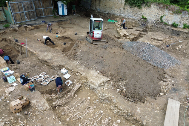 Finální vyzvednutí (exkavace) kosterních ostatků z liniového hrobu č. 12 a 17 a dvou skupinových u vjezdu na pozemek. Vlevo dole ukládání kosterních ostatků na podnosy a jejich popis antropoložkou (foto Egon Ditmar, 9. 6. 2025).